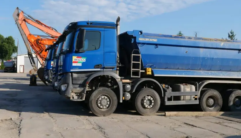 Niebieskie wywrotki i koparka stojące na placu budowy.